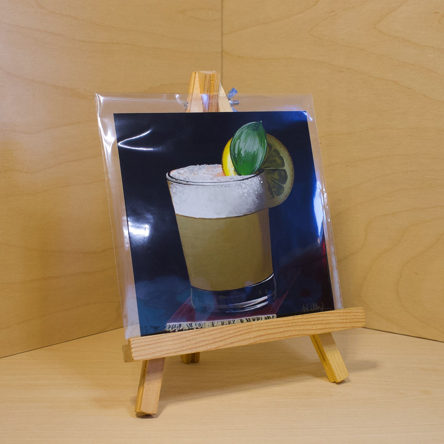 A 6x6" fine art print of the original acrylic painting "Whiskey Sour" by Hannah Kilby from Hannah Michelle Studios. Displayed in a protective plastic sleeve on a small, wooden easel.