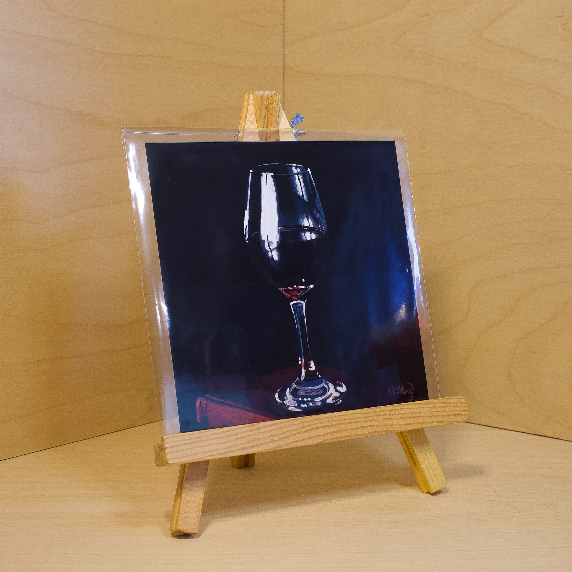 A 6x6" fine art print of the original acrylic painting "Red Wine" by Hannah Kilby from Hannah Michelle Studios. Displayed in a protective plastic sleeve on a small, wooden easel.