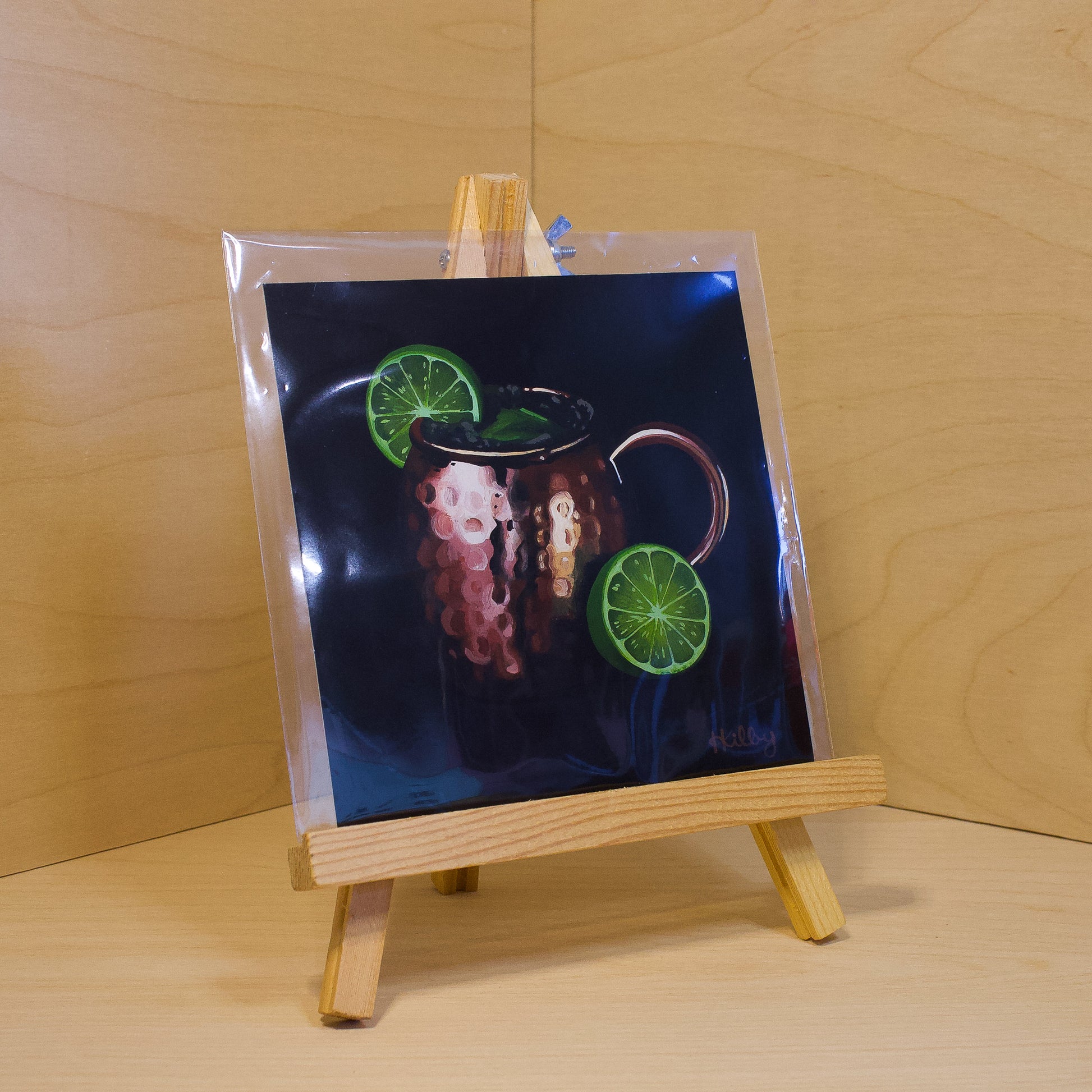 A 6x6" fine art print of the original acrylic painting "Moscow Mule" by Hannah Kilby from Hannah Michelle Studios. Displayed in a protective plastic sleeve on a small, wooden easel.