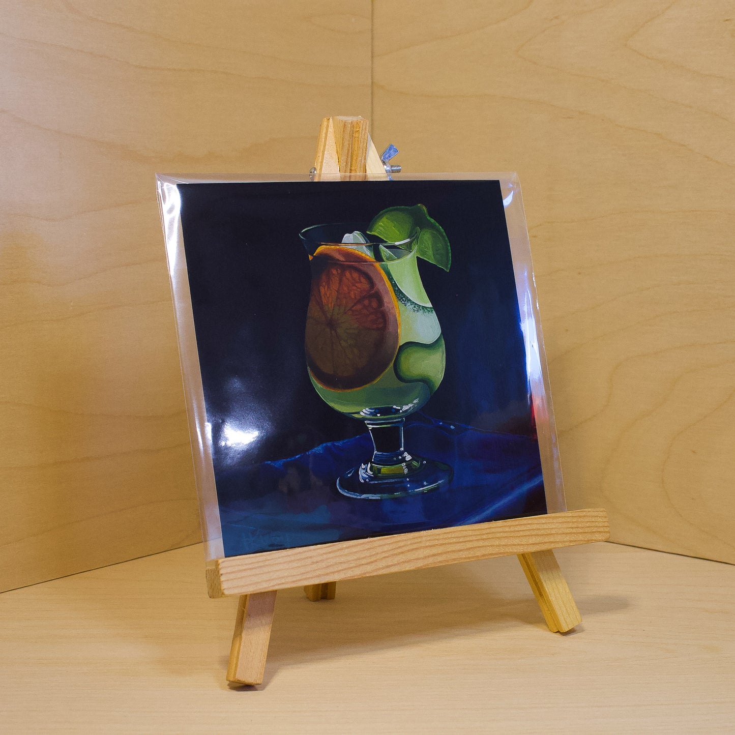 A 6x6" fine art print of the original acrylic painting "Gin & Tonic" by Hannah Kilby from Hannah Michelle Studios. Displayed in a protective plastic sleeve on a small, wooden easel.