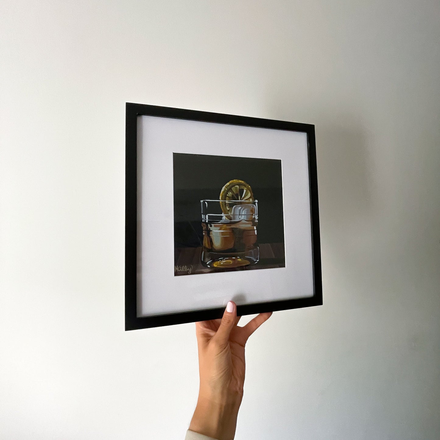 A hand holds aloft a framed 8x8" fine art print of an original acrylic painting of an Old Fashioned cocktail. The print sits in a wide, white matte and a sleek, black frame.
