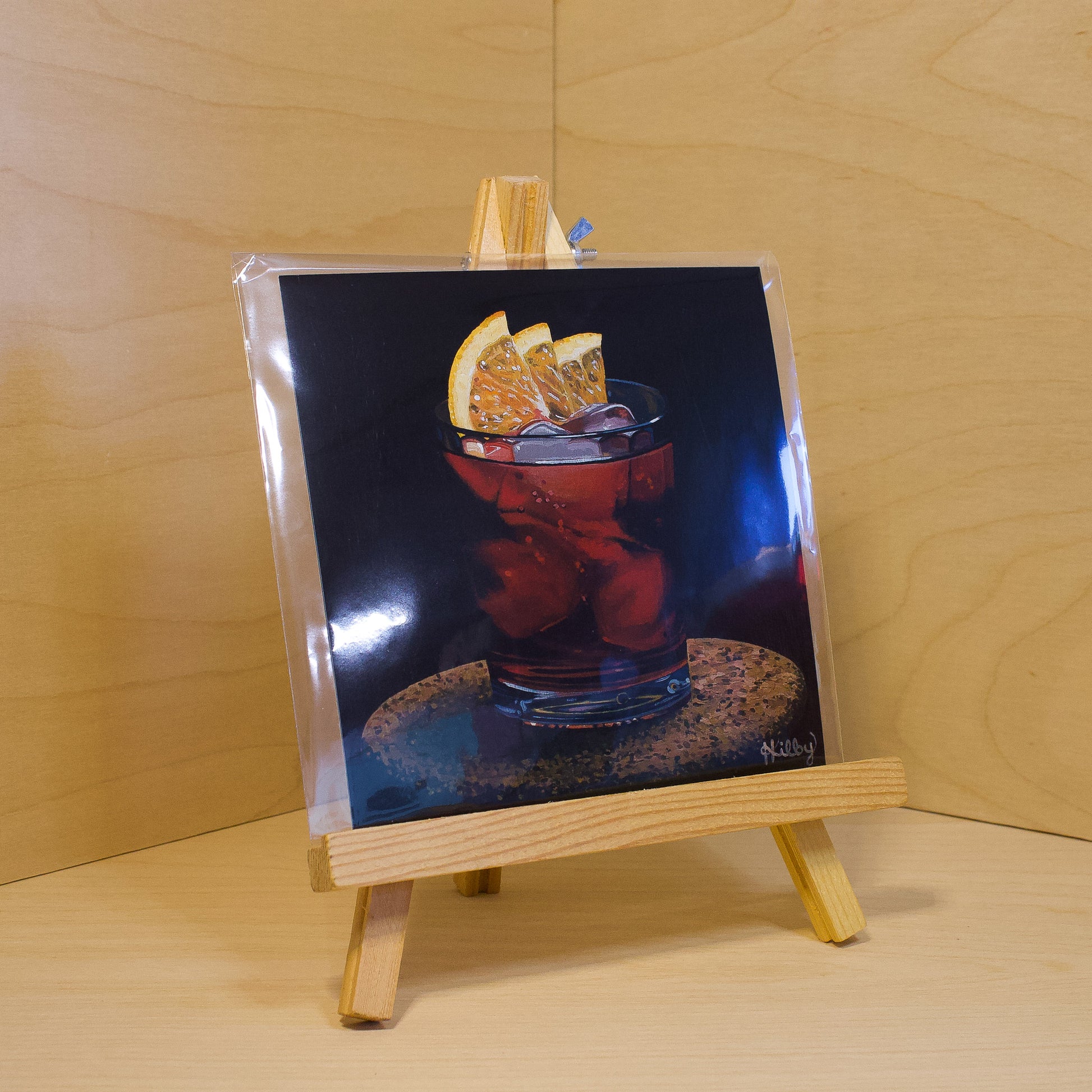 A 6x6" fine art print of the original acrylic painting "Negroni" by Hannah Kilby from Hannah Michelle Studios. Displayed in a protective plastic sleeve on a small, wooden easel.